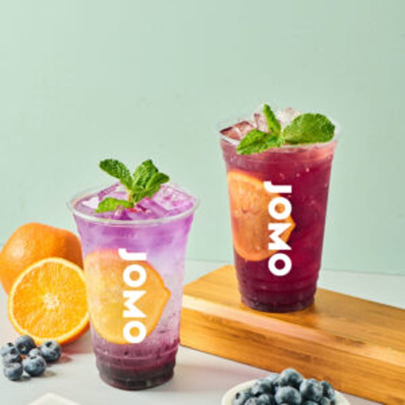 Two JOMO brand fruit drinks, one purple and one red, garnished with mint and orange slices.