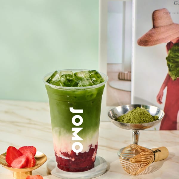 A JOMO matcha strawberry drink with ice, matcha powder, whisk, and fresh strawberries on a marble table.