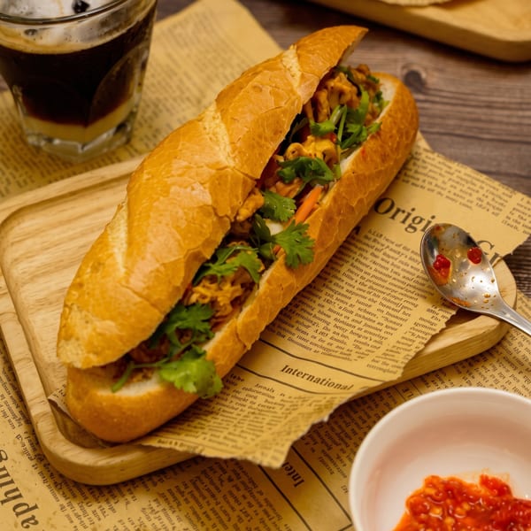 A Vietnamese Banh Mi sandwich, iced coffee, and chili sauce on a wooden board and newspaper.