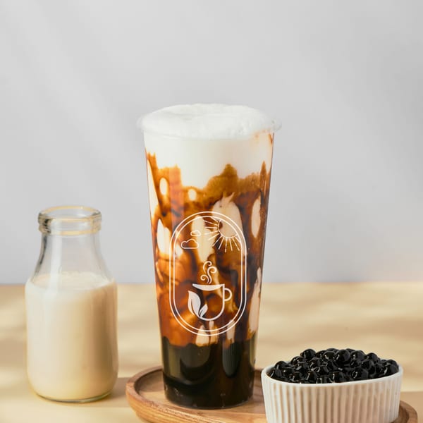 Boba tea with milk foam, a bottle of milk, and a bowl of boba pearls on a wooden tray.